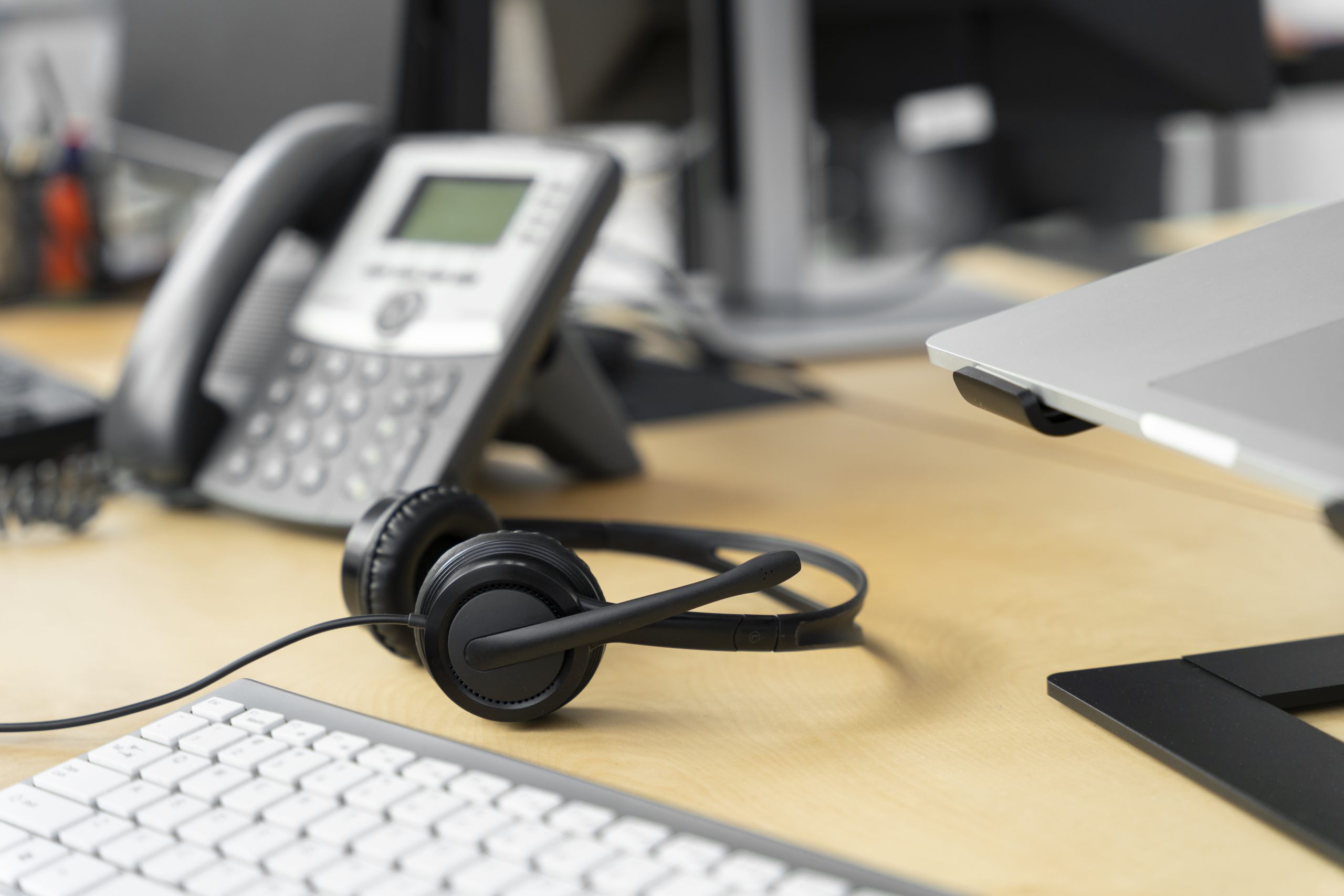 Call center headset placed on office desk with IP phone, keyboard, and laptop, representing customer support and VoIP communication services.